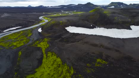 aerial drone footage of iceland dark brown and black volcanic rocky terrain landscapes during a cloudy day with a river flowing into the distance and large mountains with green moss, plants and snow.