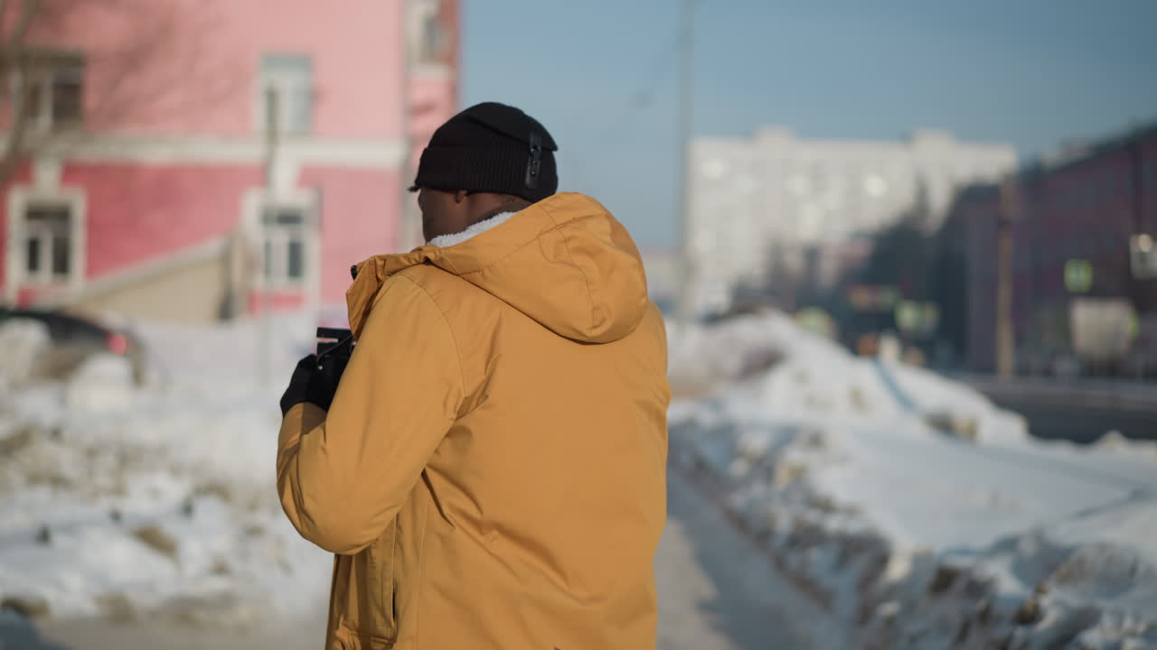 back view of dark skinned cameraman taking shot while walking along snow covered urban walkway with passing cars, exhaling visible breath into crisp frosty morning air amid city buildings