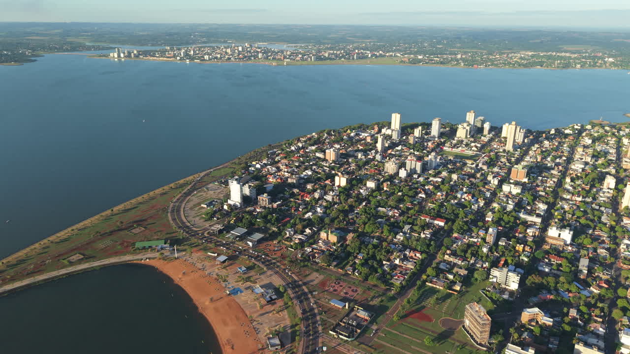 Aerial drone orbit of Posadas, Misiones, highlights El Brete beach, Paraná River, city skyline, and across distant Encarnación, Paraguay,with prominent urban and waterfront geography, real time.