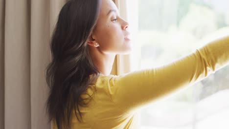 Vista-Lateral-De-Una-Mujer-Birracial-Revelando-Cortinas-Y-Mirando-Por-La-Ventana