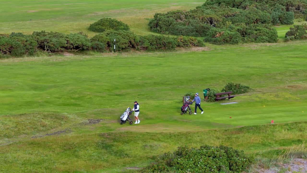 Two golfers walk with bags on a green fairway, natural daylight, wide landscape view