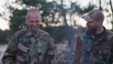 soldiers in camouflage clothing, discussing in the evening at a common bonfire frying sausages for supper, men joking, laughing, resting after work, service, field exercises