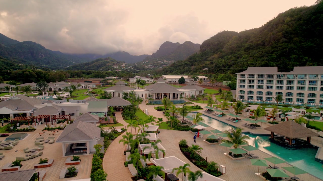 Una fluida ripresa aerea con drone sopra il complesso centrale di piscine di un lussuoso resort caraibico, che rivela l'architettura delle ville, i lussureggianti giardini con palme e uno spettacolare sfondo montuoso.