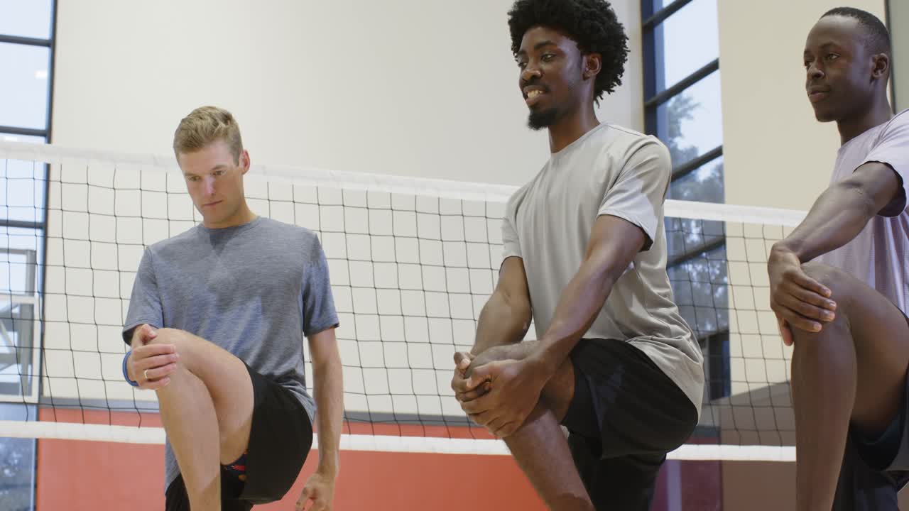 Diverse männliche Teamkollegen machen in der Turnhalle vor dem Training eine Knie-Dehnübung, Volleyballnetz sichtbar.