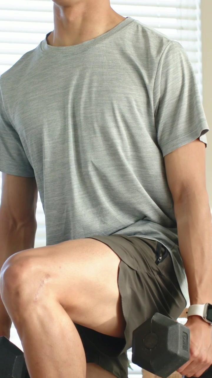 Video verticale: Uomo con maglietta grigia che solleva un ginocchio e fa affondi a casa, con manubri per il fitness.