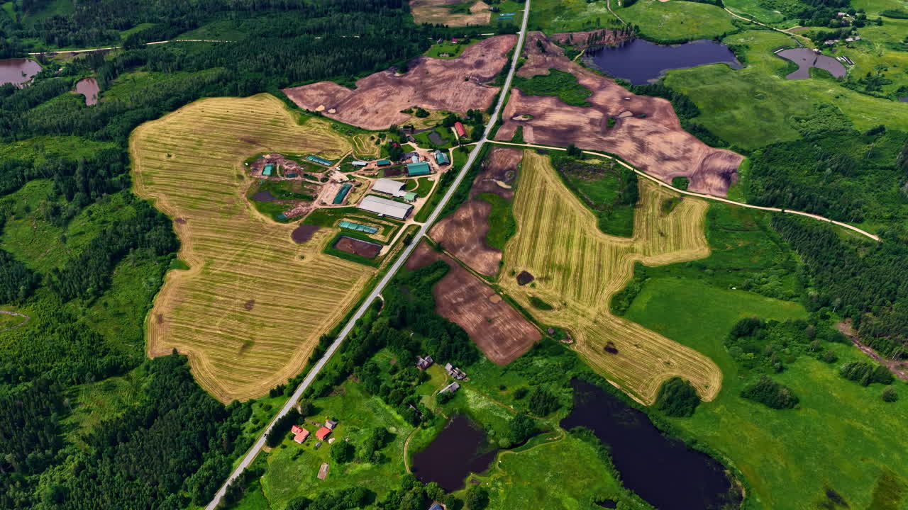 Drone aerial view of farmland, green meadows, ponds, scattered houses and a countryside road intersecting rural landscapes in Latvia during summer