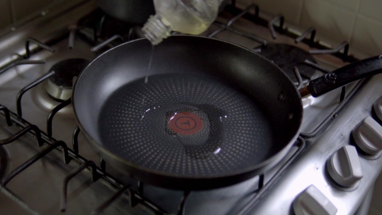 Close-up shot of oil being poured into frying pan on stove during cooking preparation.