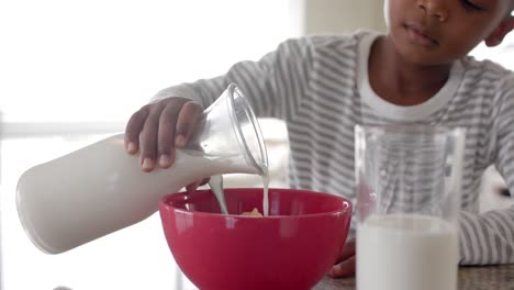african american boy having breakfast pouring milk on cereal in kitchen, slow motion