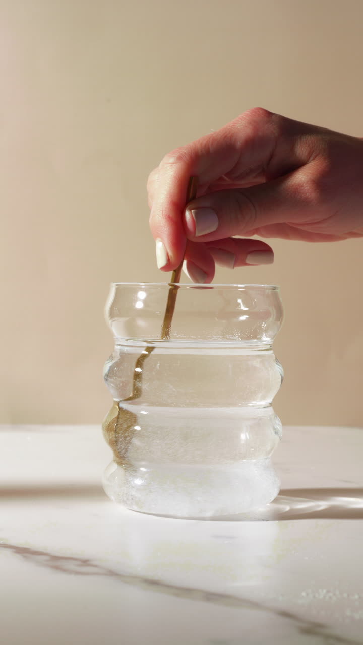 Close-up of a hand stirring powder, resembling magnesium, electrolytes, creatine, protein supplements, into water using a spoon. The powder swirls visibly in a curved glass on a white marble surface.