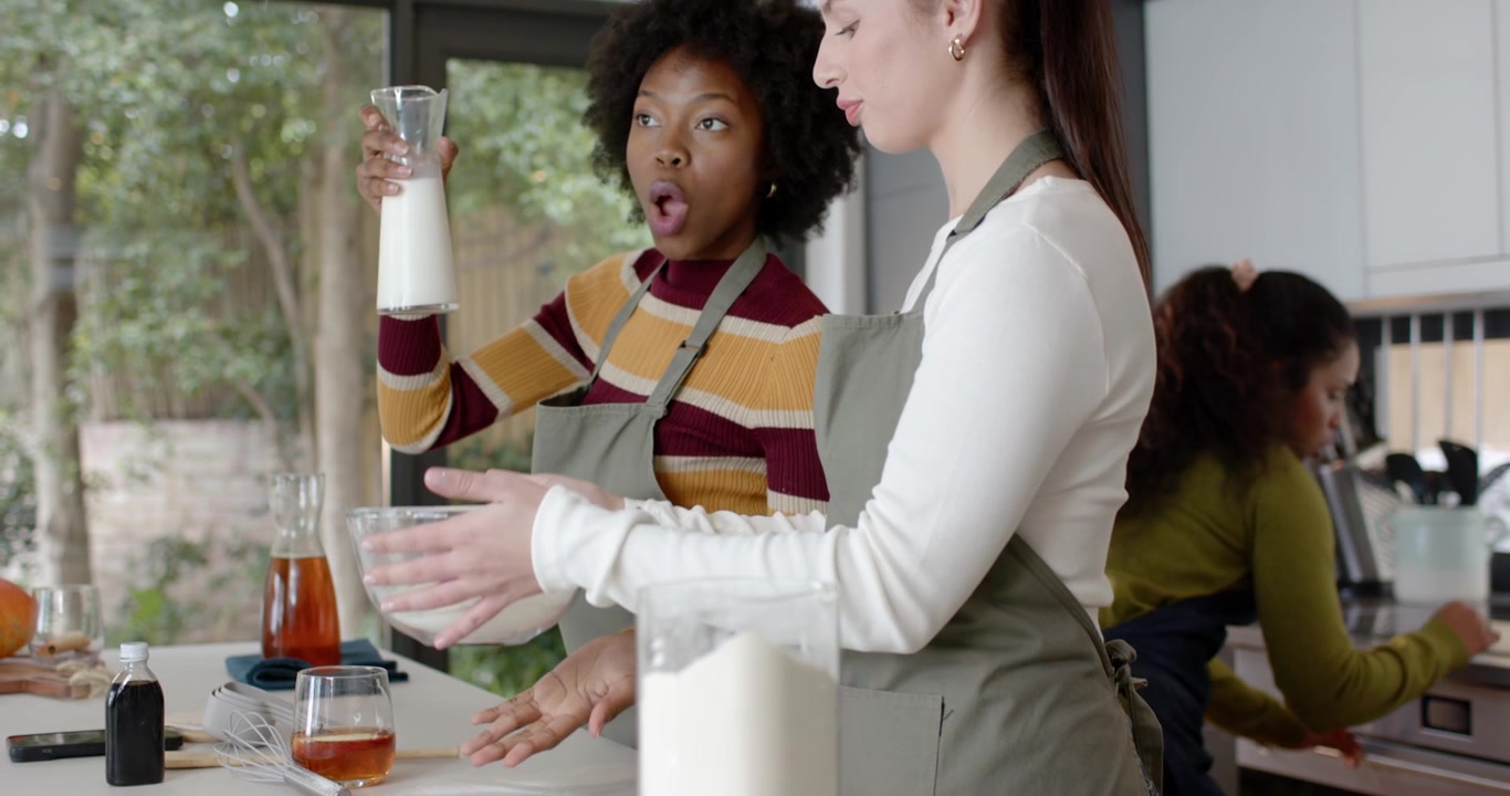 Diverse female friends in aprons reaching for milk, holding bowl, whisk at kitchen counter to bake