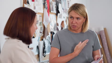 female tailor demonstrating sewing technique to student using hand gesture on chest side during learning session in creative fashion studio with materials papers sketches dummy in background