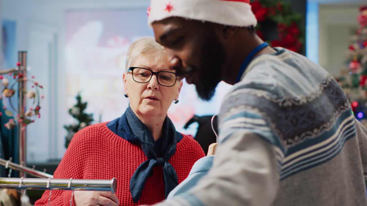 empleado afroamericano con sombrero de papá noel en una tienda de ropa decorada con navidad, ofreciendo consejos de compras a clientes mayores indecisos durante la temporada de vacaciones de navidad