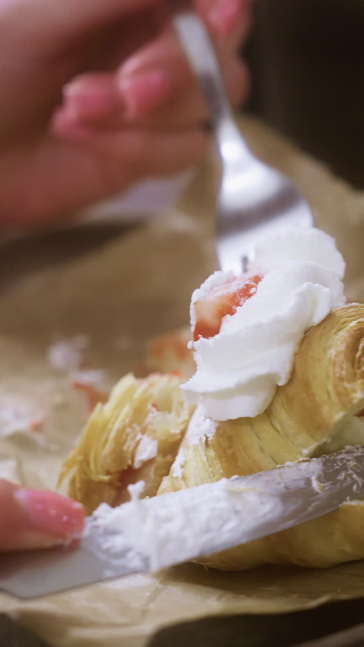 Close-up shot of female hand using fork to dig into creamy croissant topped with whipped cream and fruit. Croissant cut into with knife in cozy cafe, blurred background