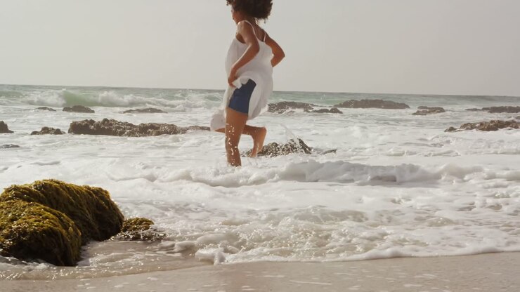 Vista laterale di donna afroamericana che gioca con l'acqua del mare sulla spiaggia 4k