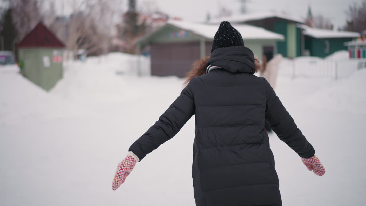 Girl in black winter jacket and knitted hat walks outdoors on snowy day, curly hair visible under hat, scarf wrapped around neck, back view showing solitude and calm moment in residential snowy neighborhood