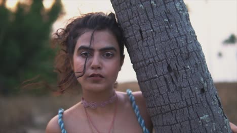 A-young-girl-in-blue-dress-holding-tree-trunk-at-the-beach-and-looking-at-the-camera