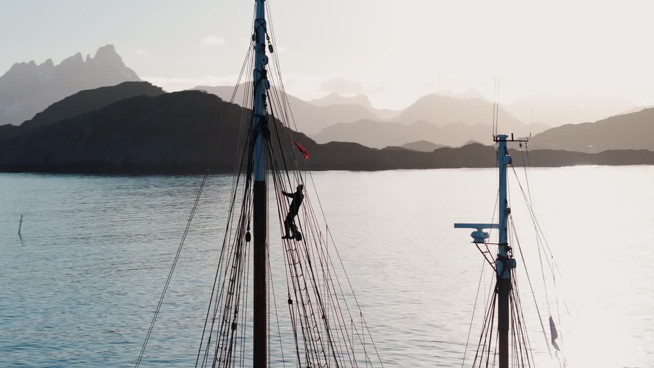 Droneoptagelse af sømand øverst i masten med udsigt over solnedgang i Lofoten, silhuet mod gylden arktisk himmel og rolig fjord, koncept for eventyr og frihed.