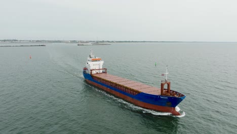 aerial establishing view of large blue cargo ship leaving port of liepaja , karosta bridge, slight overcast day, calm baltic sea, city in background, wide ascending drone shot moving forward