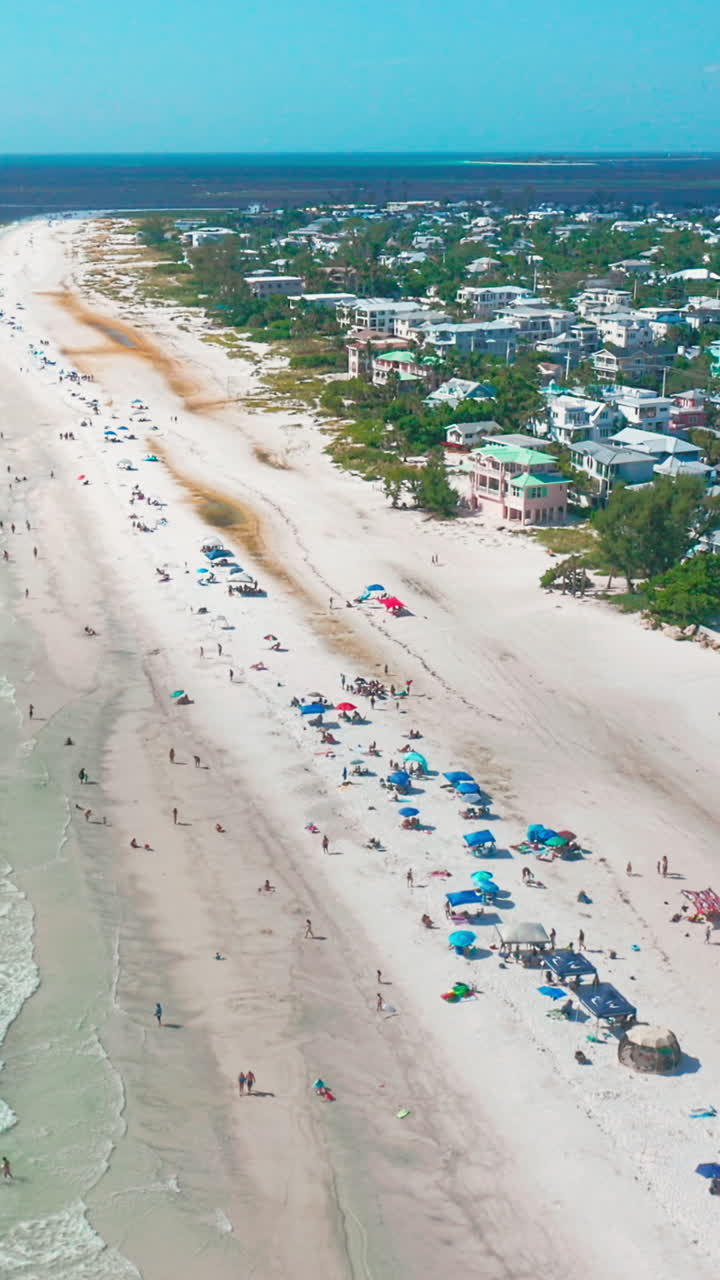 Groups of beachgoers gather beneath colorful umbrellas along a broad sandy shoreline, with turquoise waves lapping beside a quiet coastal community