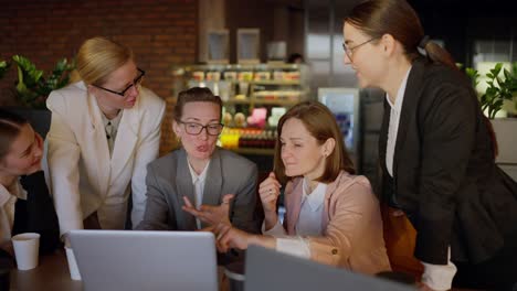 A-group-of-confident-businesswomen-in-a-business-uniform-gathered-around-a-laptop-and-a-girl-speaker-in-a-gray-suit-presenting-their-work-and-ideas-while-working-in-a-modern-office