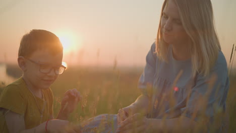 a woman in a blue gown and a young boy in a yellow shirt are sitting together in a grassy field during sunset, gently plucking and examining blades of grass, capturing a tender, peaceful moment
