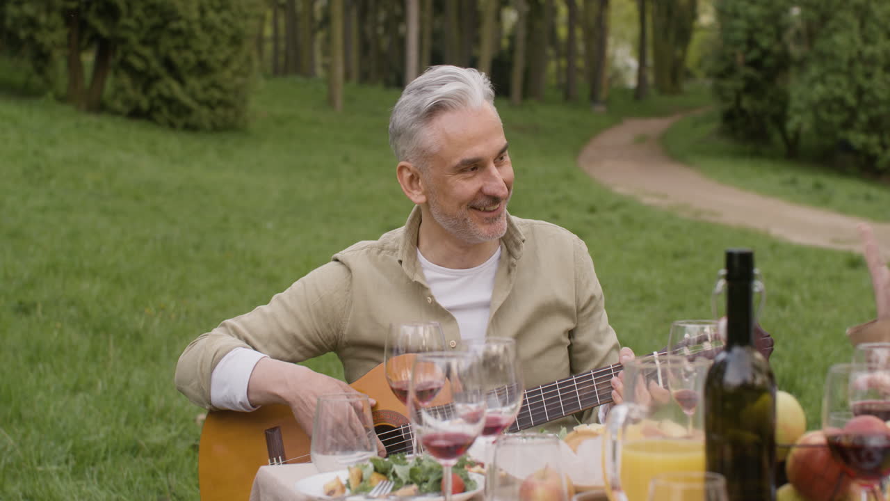 Middle Aged Man Plating A Guitar Sitting At Table With His Friends During An Outdoor Party In The Park