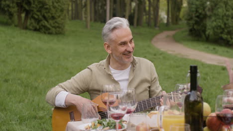 middle aged man plating a guitar sitting at table with his friends during an outdoor party in the park