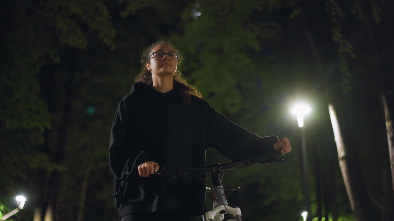 Night Rider Pauses, Woman On Bike Gazes Upward, Cyclist Contemplates Under Bright Streetlamp, Nighttime Cyclist With Thoughtful Expression Pauses To Look Into Dark Evening Sky
