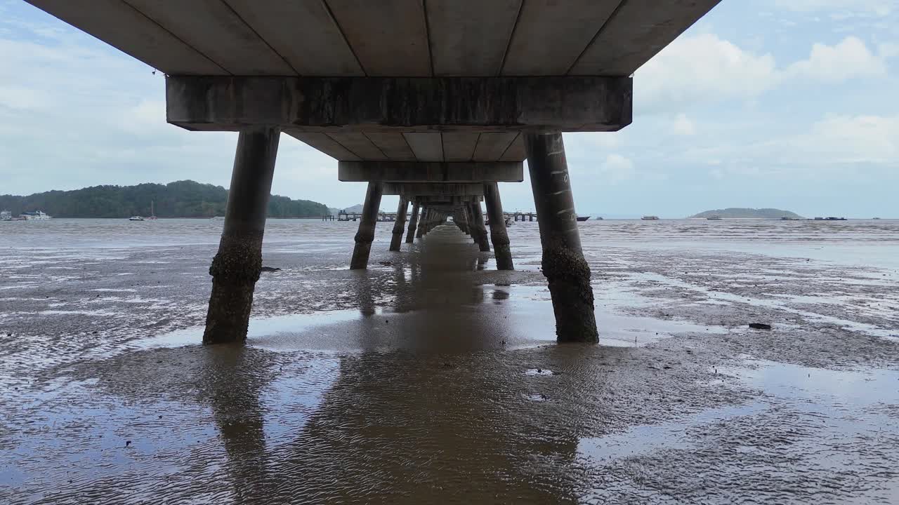 Aerial view under a pier in Phuket, Thailand, showcasing wet sand and receding tides under overcast skies