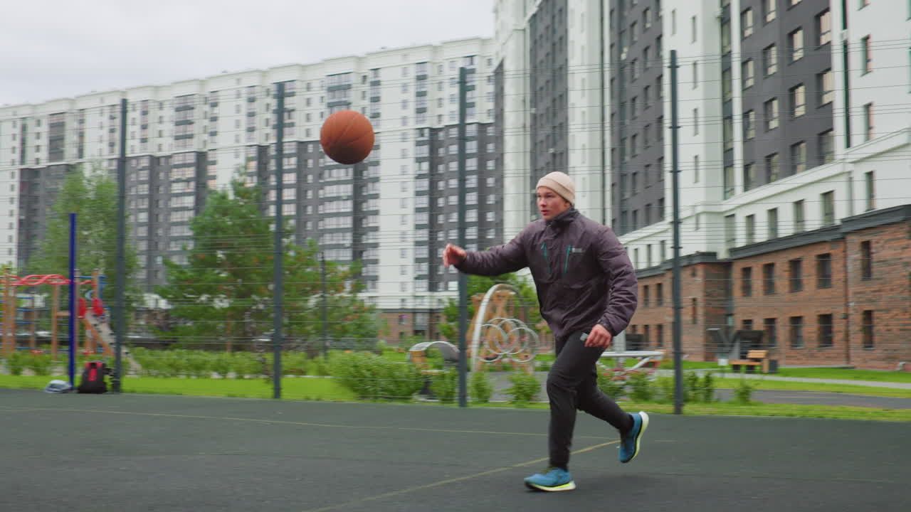 Pria kulit putih lanjut usia mendribel bola basket di lapangan perkotaan, mengenakan beanie dan jaket, berlatih layup dan jump shot di bawah langit mendung, dikelilingi oleh menara tempat tinggal bertingkat tinggi dan berpagar