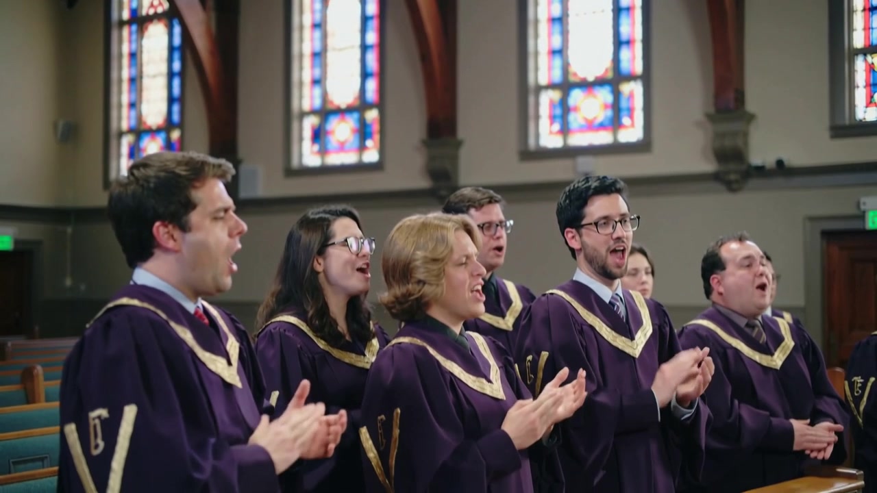 Telecamera che si avvicina a un coro in abiti viola che canta e applaude al primo banco per il culto, con vetrate colorate.