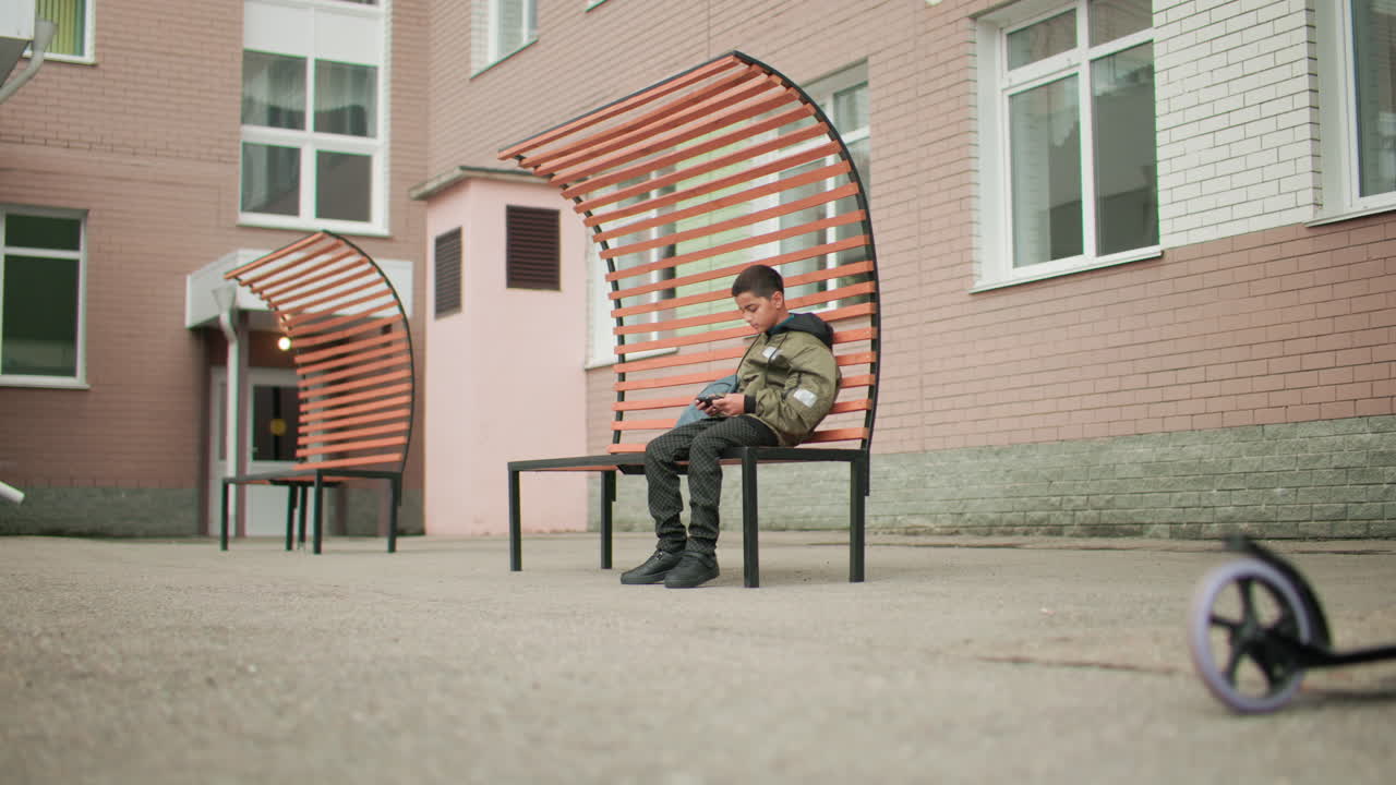 학교 쉬는 시간에 현대적인 벤치에 앉아 휴대폰 게임에 집중하고 있는 어린 학생, 옆에는 가방이 놓여 있고, 교육 건물 안뜰 근처 전경에는 스쿠터가 주차되어 있어요.