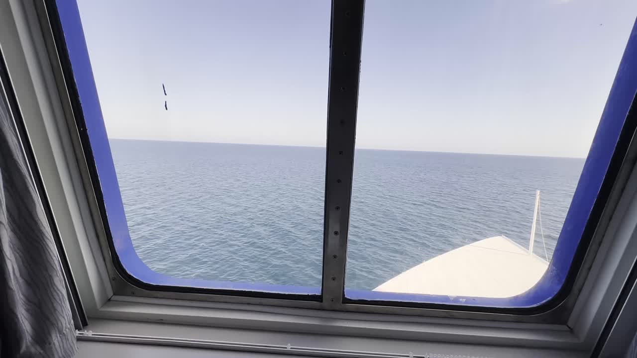 View from inside a ferry or cruise ship looking out through a window toward the open ocean. The horizon is visible in the distance, along with the front edge of the vessel cutting through calm waters