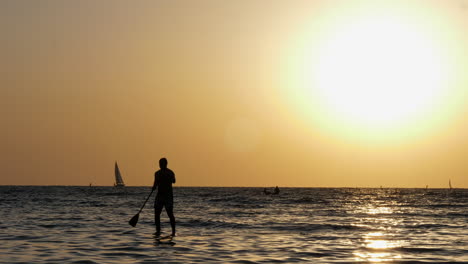 man in silhouette stand-up paddling in the sea against golden sun in the sky in tel aviv, israel