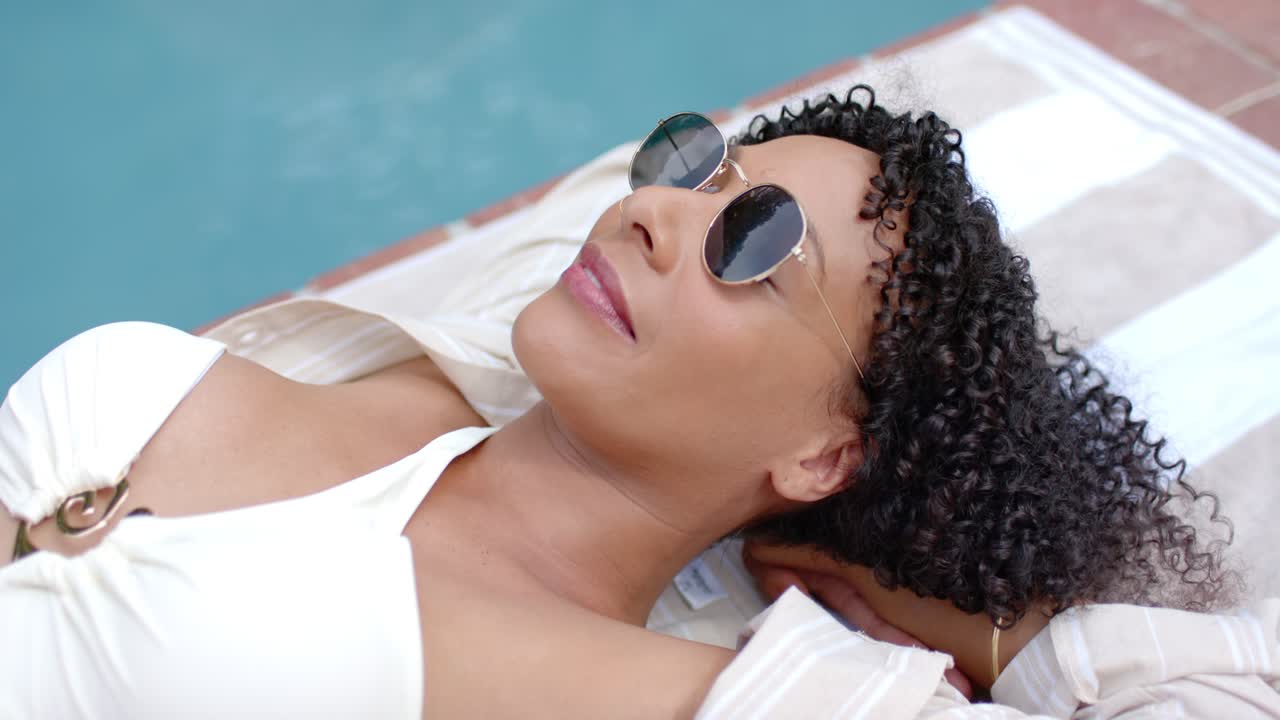 Woman reclining, relaxing in white swimsuit on towel by pool, camera panning sunglasses reflecting
