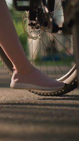 Side view of individual legs in pink sneakers kicking bicycle tire to check air pressure, lush greenery and trees create beautiful backdrop, with shadow details and car passing in blurred background
