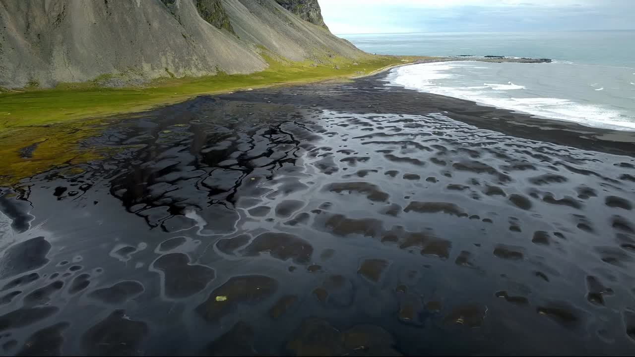 Drone shot of winding dirt road passing through green marshland near dark mountains in Iceland. Scenic route follows edge of water with dramatic rocky peaks rising in background under soft light.