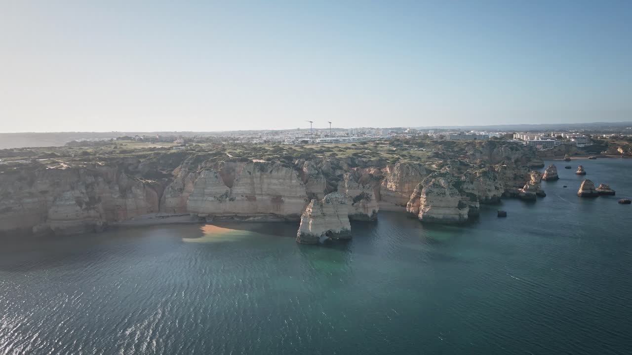 Camera spirals along sunlit cliffs and turquoise waters, revealing the intricate coastline, rugged beaches, and distant Portuguese town