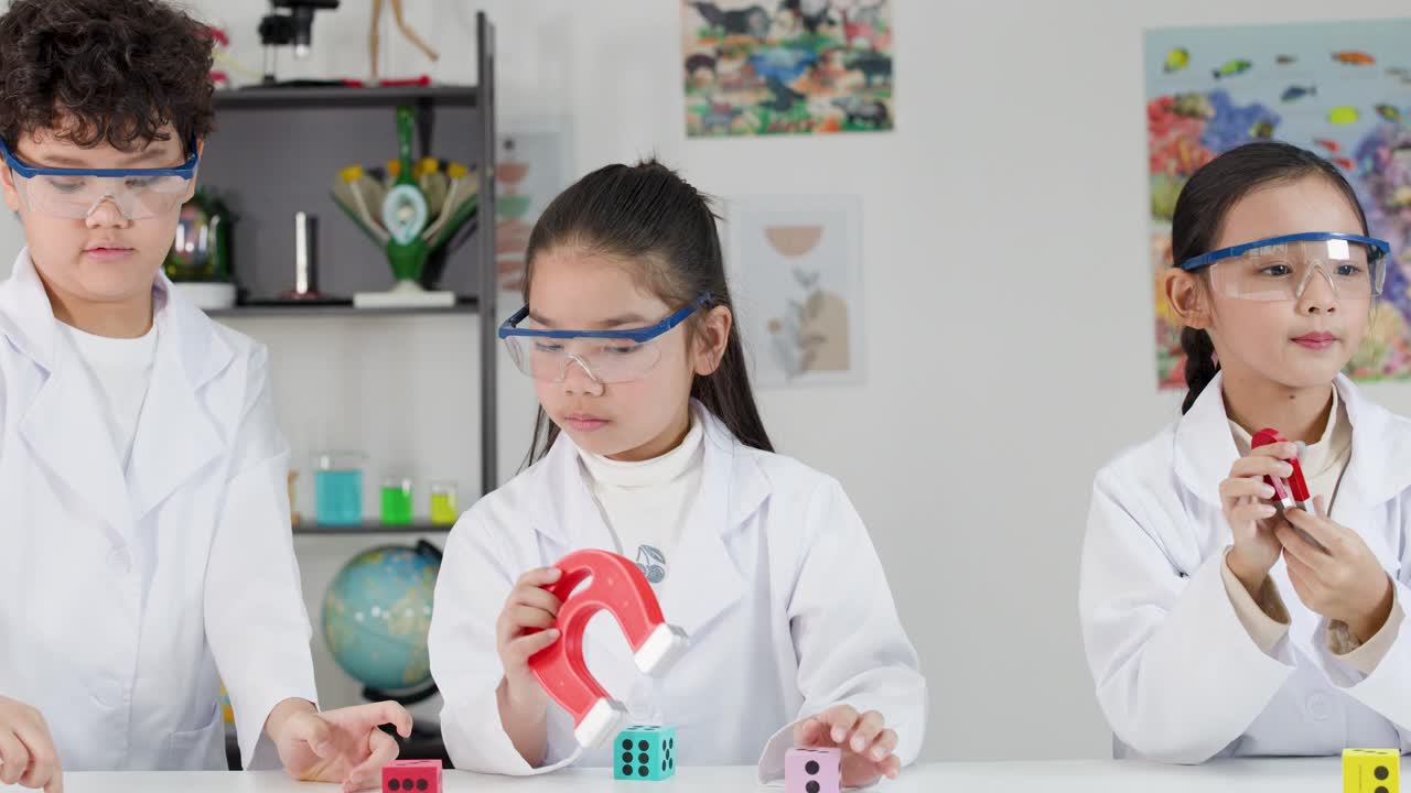 Tiga siswa muda mengenakan jas lab dan kacamata pelindung saat mempelajari magnetisme di ruang kelas yang cerah.