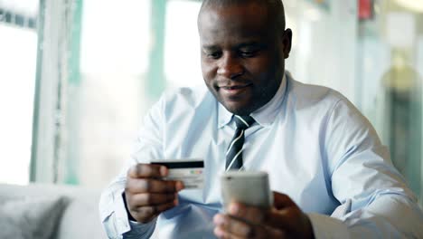 cheery african american businessman in formal clothes paying online bill keeping credit card and smartphone in his hands in glassy cafe