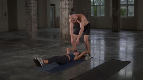 active young woman is exercising abdominal muscles training with trainer, lifting legs and body lying on mat, man force it back. people and sports concept. loft studio. slow motion