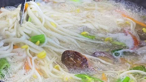 Almeja-De-Marisco-Kalguksu-Hirviendo-En-Una-Olla---Pinzas-Mezclan-Sopa-De-Fideos-Cortada-Con-Cuchillo-Mientras-Se-Cocina-Primer-Plano