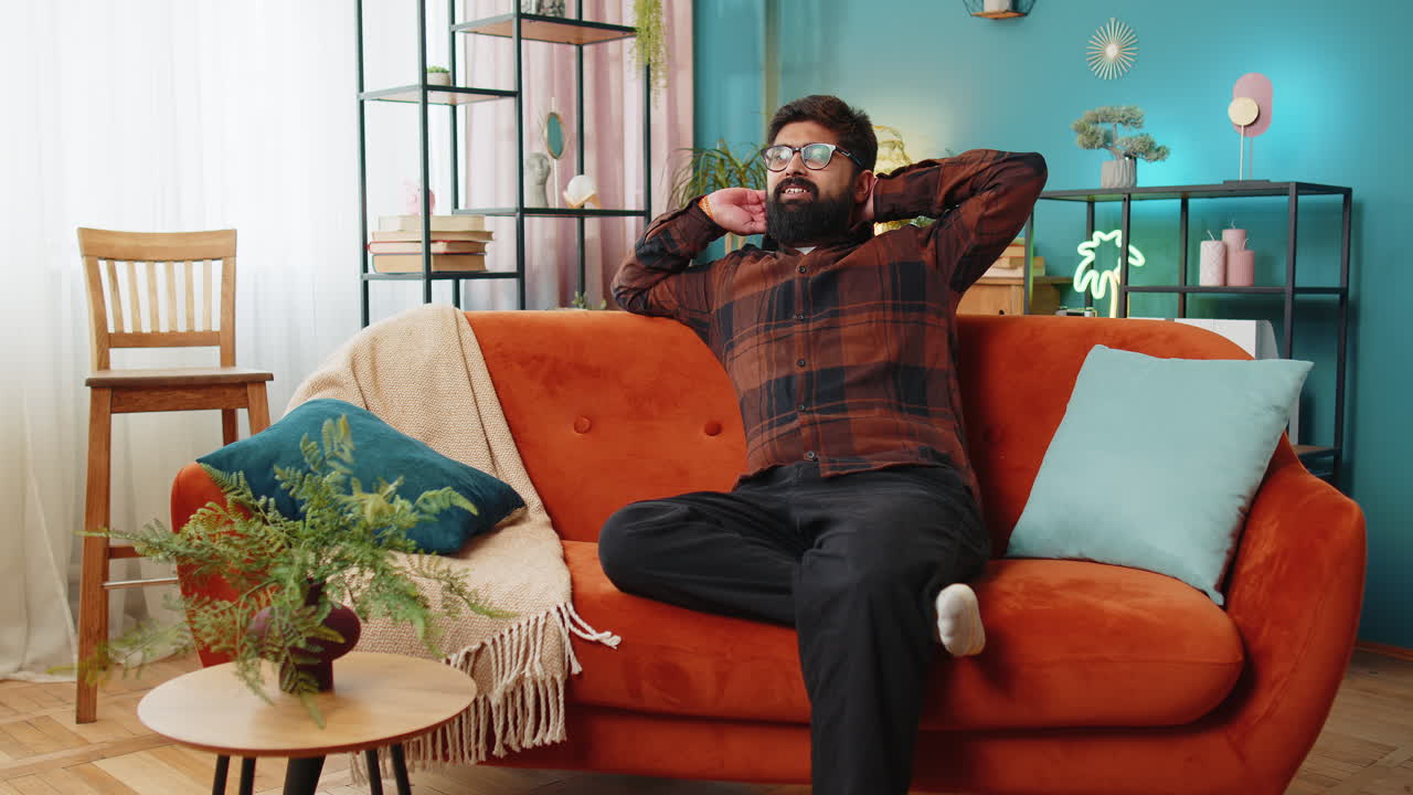 Indian man entering living room relaxing taking a break resting puts hands behind head on sofa