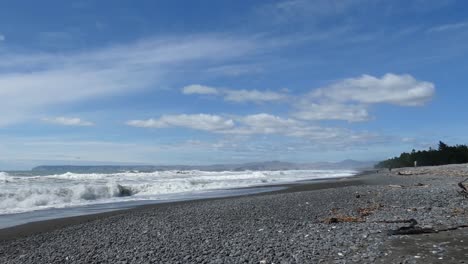 Menschen-Laufen-Am-Steinigen-Strand-Entlang,-Während-Hunde-In-Die-Wildwasserbrandung-Laufen-–-Rarangi-Beach,-Marlborough