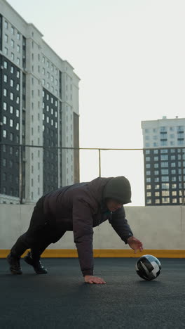atleta realizando flexiones dinámicas alternando la colocación de la mano en una pelota de fútbol demostrando fuerza, coordinación y agilidad contra un telón de fondo de postes de gol y edificios residenciales