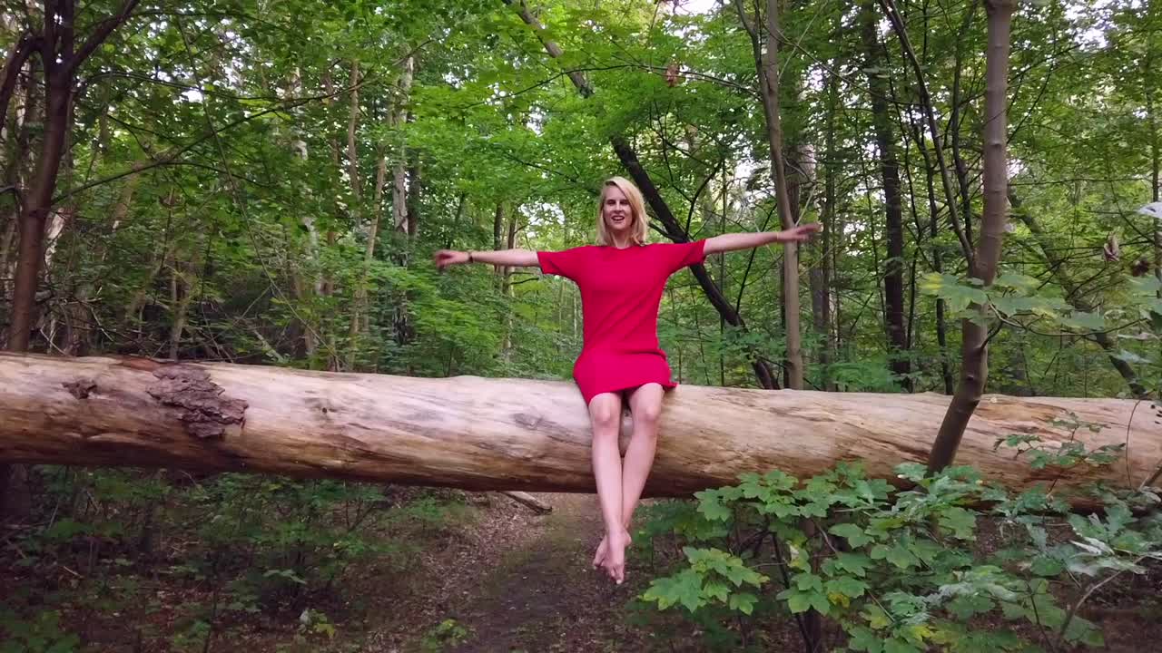 Perfect slow motion cinematic shot of a lonely blond barefoot girl in a red dress sitting alone on a big tree in the forest. Foto video shoot on lost places in nature 1080, 24p by Philipp Marnitz