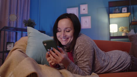 mujer acostada en el sofá en casa usando el teléfono móvil comunicándose en redes sociales o compras en línea