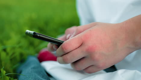 close-up of hand holding smartphone and operating it while seated on grassy field, surrounded by lush greenery, focus on fingers and phone screen with a serene outdoor atmosphere