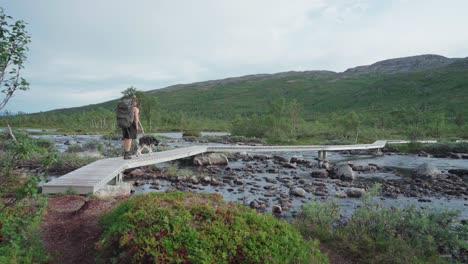 caucasian guy hiking with a husky dog crossing a rocky river on wooden footpath at anderdalen national park in senja, norway