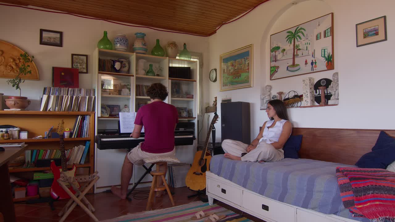 A musician playing the keyboard in his home studio in Capri while his girlfriend watches.